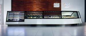 A modern, curved glass display case filled with various desserts and pastries stands in a sleek, minimalist room with white floors, a wooden accent wall, and geometric hanging lights above.