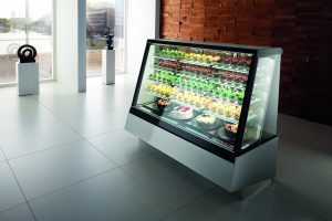 A modern glass display case in a bright, minimalist room showcasing assorted colorful pastries and cakes on multiple shelves. A wooden accent wall and large windows are in the background.