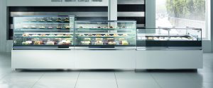 A modern pastry display case in a bright, clean bakery, filled with assorted cakes, pastries, and desserts arranged on glass shelves. Large windows reveal a sunlit outdoor scene with greenery.