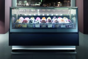 A glass display case filled with assorted colorful gelato flavors, each in separate bins with decorative toppings, stands in a modern indoor setting with city lights visible through the window behind it.