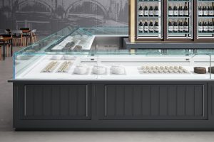 A modern bakery display case with glass panels showing cakes, pastries, and desserts inside. Wine bottles are visible in a fridge behind the counter. The setting is clean and contemporary with gray décor.