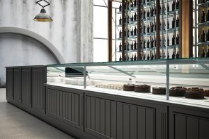 A modern bakery display case with several chocolate cakes and desserts, situated in a sleek, industrial-style interior with a wall of wine bottles in the background and a large, round window to the left.