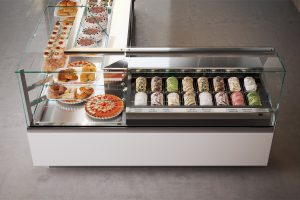 A modern glass display case filled with assorted gelato flavors in metal trays, alongside another case displaying assorted pastries and desserts on plates. The counter is clean and minimalist, set on a gray floor.