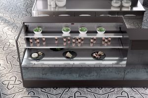 A display case in a modern kitchen shows bowls of leafy greens on the top shelf, small bottles in the middle, and assorted cheese and meats on black plates on the bottom shelf. The floor has a geometric patterned tile.