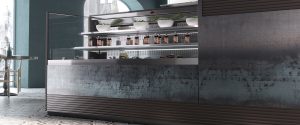 A modern café counter with glass panels displays bowls of fresh greens on top shelves and bottles of sauces or dressings on the middle shelf, with a dark, textured front and patterned tile floor.