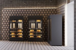 Two black display refrigerators filled with large wheels of cheese stand against a hexagonal patterned wall in a modern, tiled room, with a third closed refrigerator on the right.