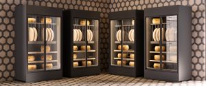Four black display refrigerators with glass doors show large wheels of cheese on shelves inside, set against a wall and floor with a hexagonal tile pattern.