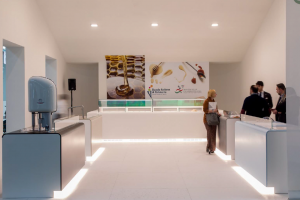 A modern, minimalist exhibition booth with white counters and display cases, featuring large food-themed posters on the back wall. Several people are standing and conversing on the right side of the booth.