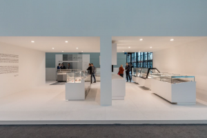 A modern, well-lit museum exhibition space with several glass display cases. People are viewing artifacts inside the cases. Large windows are visible in the background on the right side.