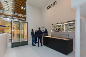 Five men in business attire stand and talk inside a modern exhibition booth with glass display cases, a wall of bottles, and a vertical rotisserie, under a sign that reads CIAM.
