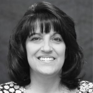 Black and white portrait of a woman with shoulder-length dark hair and bangs, smiling at the camera. She is wearing a patterned top, and the background is plain and dark.