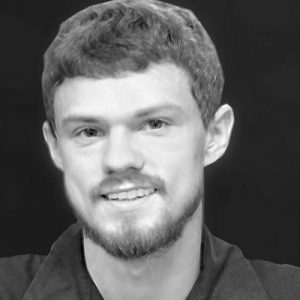 A young man with short, light brown hair and a trimmed beard is smiling. He is wearing a dark shirt and is photographed against a plain, dark background.