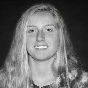 A young person with long, straight, light-colored hair is smiling at the camera. They are wearing a tie-dye shirt, and the background is dark and blurred. The photo is in black and white.