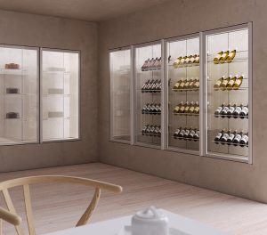 A modern wine display room with glass shelves holding various bottles of wine. There is a wooden chair and a white table with a sugar bowl in the foreground, and pastries on shelves in the background.