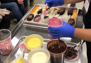A person wearing blue gloves decorates chocolate-covered ice cream bars with pink sauce and colorful sprinkles. Cups of yellow, white, and dark chocolate sauces are on a tray nearby. Others are seated in the background.