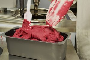 A person uses spatulas to spread vibrant pink gelato in a metal container, likely in a kitchen or gelato shop setting.