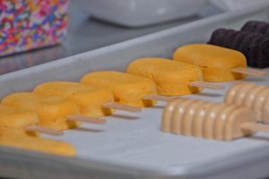 Several rows of colorful popsicles, mostly yellow-orange and tan, are lined up on a parchment-covered baking sheet. A container of colorful sprinkles is visible in the background.