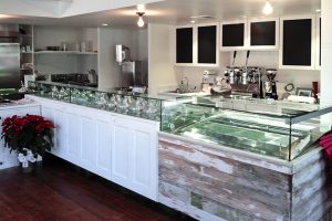 A modern, minimalist gelato shop with a glass display counter, empty metal trays, coffee machines, white cabinets, and potted poinsettia on a wooden floor.