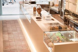 Modern ice cream shop interior with a sleek white counter, glass display cases showing a variety of fresh fruits, toppings, and pastries, with equipment and jars of cookies behind the counter. People are visible through the window.