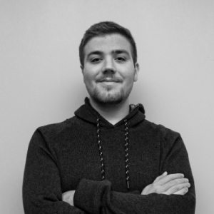 A young man with short hair and a trimmed beard stands against a plain background, wearing a dark hoodie and smiling with his arms crossed. The image is in black and white.
