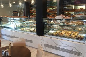 A bakery display counter filled with assorted pastries, sandwiches, and baked goods. A staff member stands behind the counter, with bread and menu boards visible in the background. Tables and chairs are in the foreground.