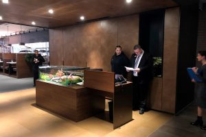 A restaurant reception area with a modern wooden desk and a display case of fresh food. Two people stand behind the desk, one reading papers. Another person stands nearby holding a blue folder. A server is in the background.
