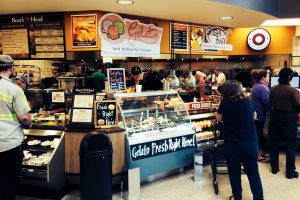 A busy supermarket deli area with gelato, sushi, and sandwich counters. Customers are ordering food, and staff are serving behind the counters. Display cases show gelato flavors, pizza slices, and other fresh foods.