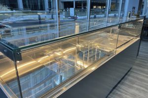 A modern, empty glass display case with internal lighting sits in front of large windows overlooking a city street on a winter day. The case has several clear shelves and compartments visible inside.