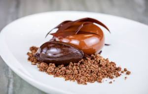 A plated chocolate dessert with a glossy dome, chocolate shards on top, a quenelle of dark chocolate mousse, and a bed of chocolate crumble on a white dish.