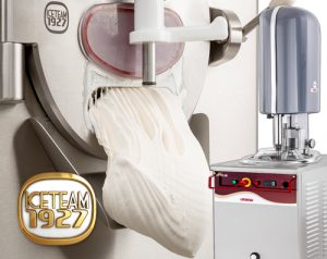 Industrial gelato machine dispensing a thick ribbon of white gelato; stainless steel equipment is visible with a logo reading ICETEAM 1927 and control panel on the right.