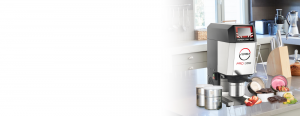 A commercial ice cream and gelato machine labeled VERSA PRO 360 sits on a kitchen counter with bowls of colorful ice cream, fruit slices, and stainless steel containers nearby. Kitchen appliances and utensils are in the background.