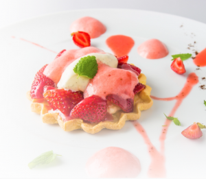 A strawberry tart topped with cream and pink sauce, garnished with a mint leaf, sits on a white plate decorated with strawberry halves, sauce dots, and mint leaves.
