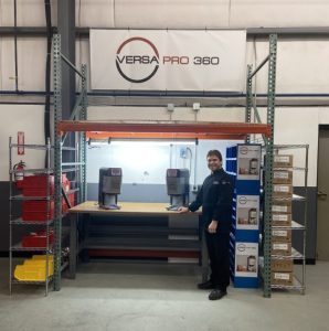 A man in a black uniform stands and smiles beside a workbench with two machines under a Versa Pro 360 banner. Shelves with red and yellow bins and stacked boxes are nearby in an organized industrial workspace.