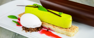 A gourmet dessert features a bright green mousse atop a rectangular biscuit, served with a scoop of white sorbet, chocolate crumble, red sauce, fresh herbs, and a curved piece of dark chocolate on a white plate.