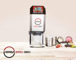 A Versa Pro 360 food machine is on a light wooden surface with metal containers, chocolate pieces, and strawberries beside it. The Versa Pro 360 logo appears in the bottom left corner.