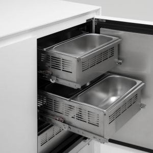 A white cabinet with its door open, revealing two stainless steel sliding drawers or trays inside, commonly used for food storage or preparation in commercial kitchens.