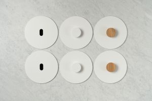 Six white round objects are arranged in two rows of three on a light marble surface. Each has a central knob: two knobs are wooden, two are plain white, and two are elongated black slots instead of knobs.