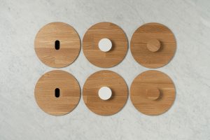 Six round wooden lids are arranged in two rows on a light marble surface. The lids have different handles: two with white knobs, two with wooden knobs, and two with elongated oval cut-outs.