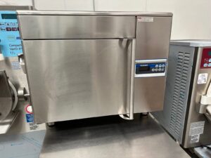 A stainless steel commercial kitchen oven with a digital control panel displaying numbers, placed on a metal countertop next to other professional kitchen equipment.