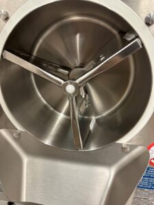 Close-up of the inside of a stainless steel industrial food mixer or ice cream machine, showing three metal mixing arms arranged in a triangular formation around a central axis.