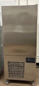 A large, stainless steel TECHFROST commercial freezer on wheels with a digital control panel and ventilation grille at the bottom, positioned against a plain wall.
