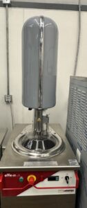 A commercial gelato machine with a large gray cylindrical lid, metal base, and control panel labeled effe 6B by cattafiga, situated in an industrial kitchen setting.