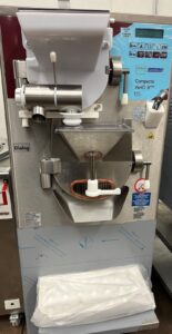 A commercial gelato or ice cream batch freezer machine with various buttons, levers, and a chute for dispensing product. A plastic-wrapped tray sits below the dispenser to collect the finished gelato or ice cream.