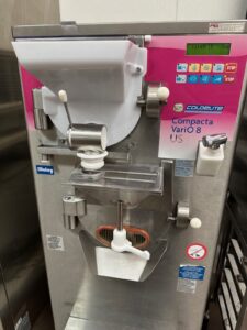 A commercial Coldelite Compacta Vario 8 batch freezer with a pink control panel, buttons, and levers, used for making gelato or ice cream, is shown in a clean industrial kitchen setting.