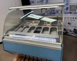 A blue commercial display freezer with a glass front, containing several empty stainless steel trays, is set indoors on a pallet with promotional posters in the background.
