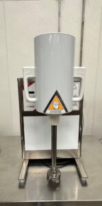 A commercial food processor with a large, cylindrical gray container, two side handles, and a warning label on the front, placed on a stainless steel countertop against a white tiled wall.