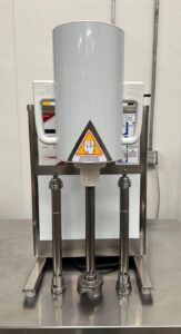 A stainless steel industrial machine with a tall cylindrical container, three vertical metal rods, and warning labels, positioned on a metal countertop in a clean, white-walled room.