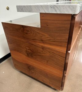 A close-up of a wooden reception desk with a marble countertop, showcasing the rich wood grain and modern design. The desk has clean lines and smooth surfaces, and sits on a reflective brown floor.