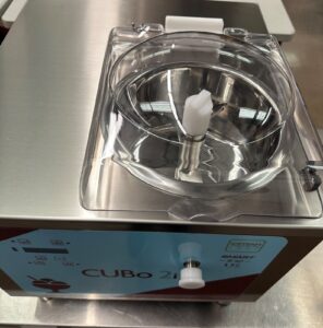A close-up of a stainless steel ice cream machine labeled CUBO 2i SMART US, with a clear lid and white knob on top. The control panel is partially visible at the front.