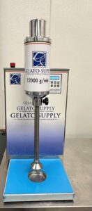 A stainless steel industrial gelato dispenser labeled Gelato Supply sits on a metal counter, featuring a digital control panel, a dispensing spout, and a sign showing a capacity of 12,000 g/min.